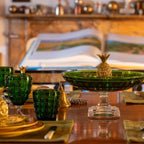 A styled dining table setting with multiple green glass goblets, gold and white plates, a large green pedestal bowl with a pineapple topper, and matching green and blue pineapple accents, photographed in a warm indoor room with a blurred book and lamp in the background.